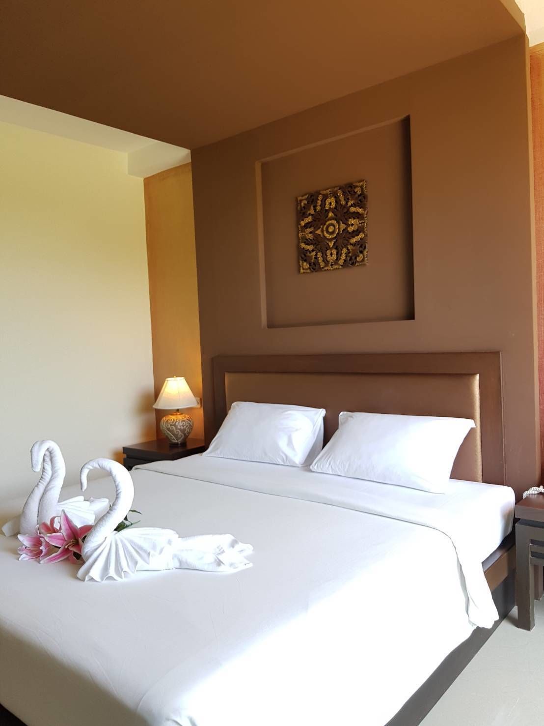 Habitación de hotel con una cama grande, ropa blanca, cabecera, una obra de arte colgada en la pared, una lámpara de noche y un arreglo decorativo de cisne y lirios sobre la cama.