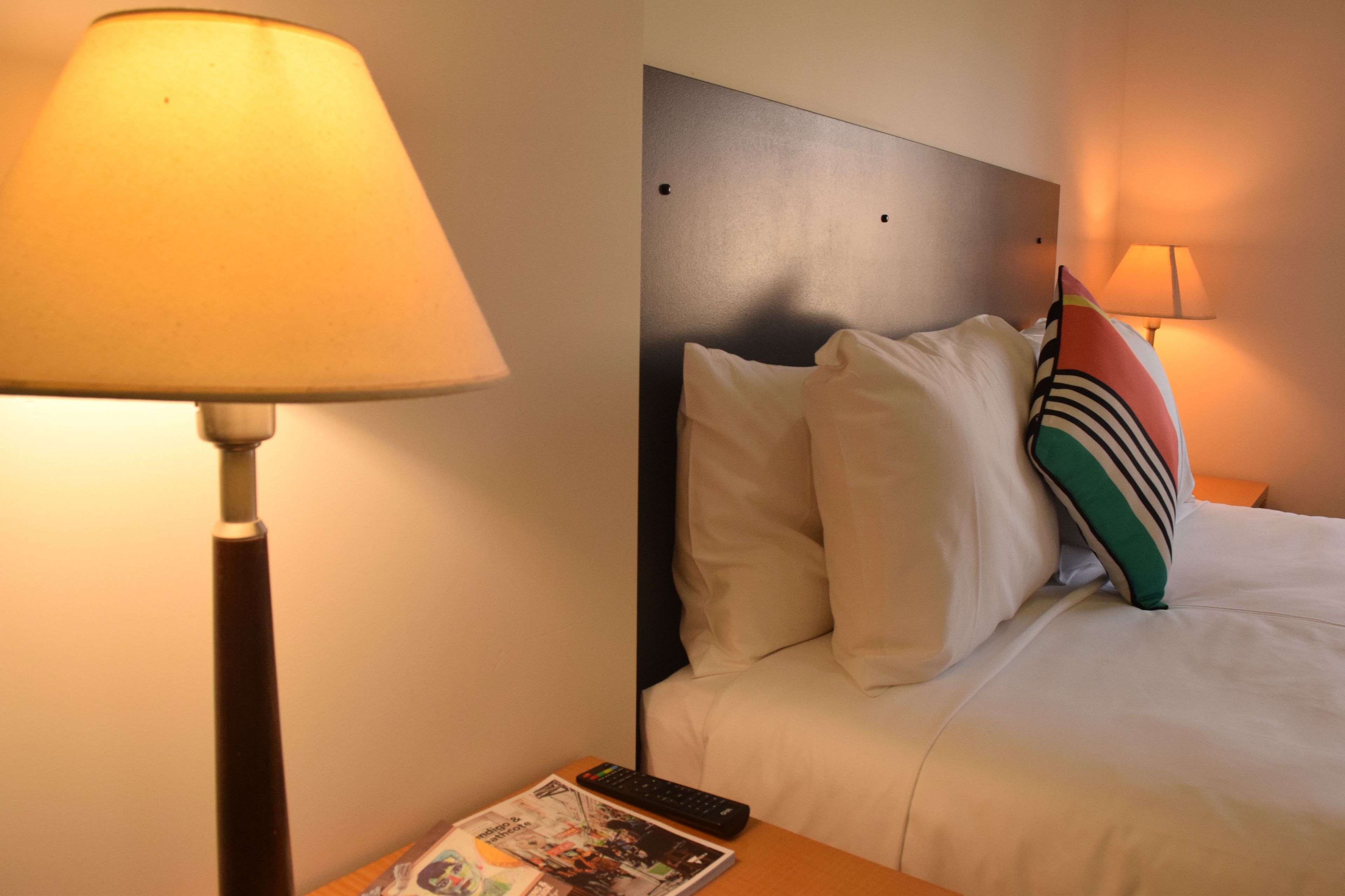 A hotel room with a bed, pillows, a remote control, and a magazine on the nightstand.