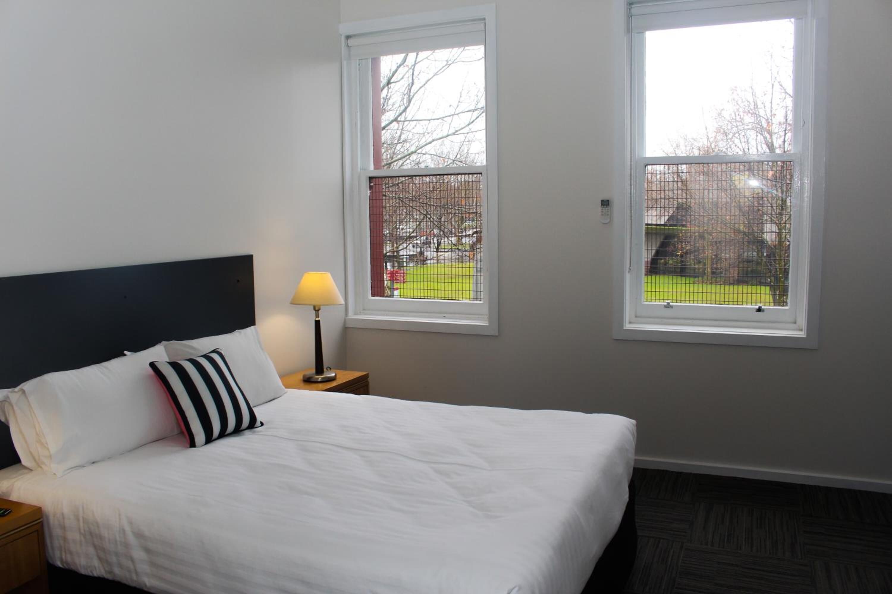 A hotel room with a bed, a nightstand, a lamp, and two windows.