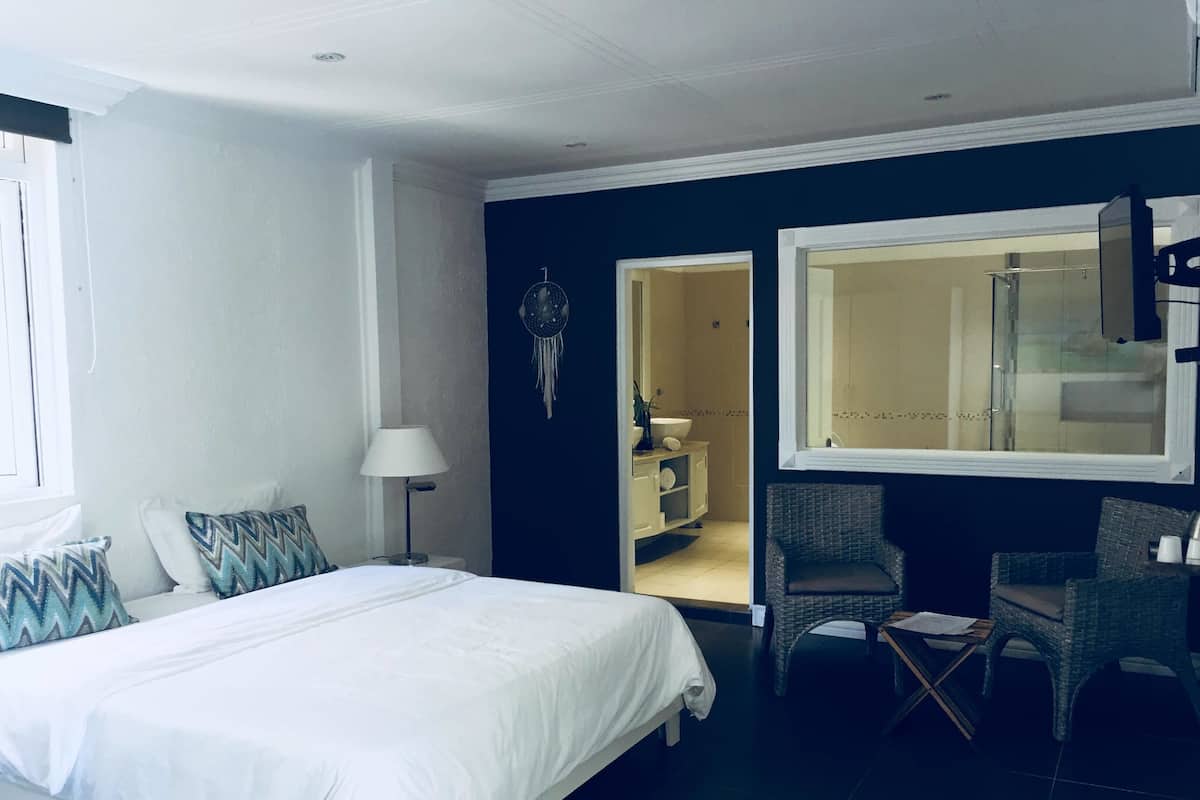 Quarto com cama, cadeira, mesa de cabeceira, luminária e banheiro visível através de uma porta aberta.