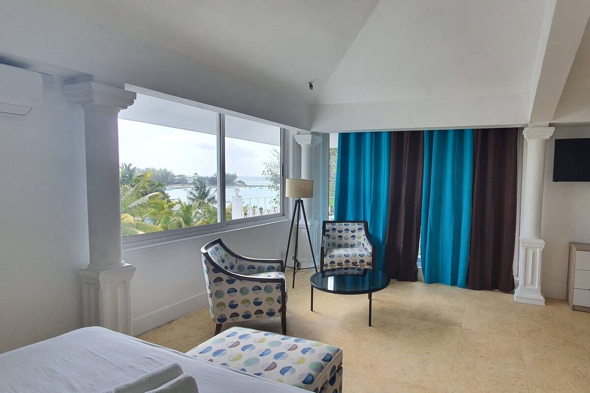 Quarto de hotel com uma janela grande, uma cama, uma cadeira, uma mesinha e uma TV fixada na parede.