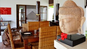 Un comedor con mobiliario de mimbre, una escultura de cabeza de Buda sobre un pedestal y una decoración floral roja.