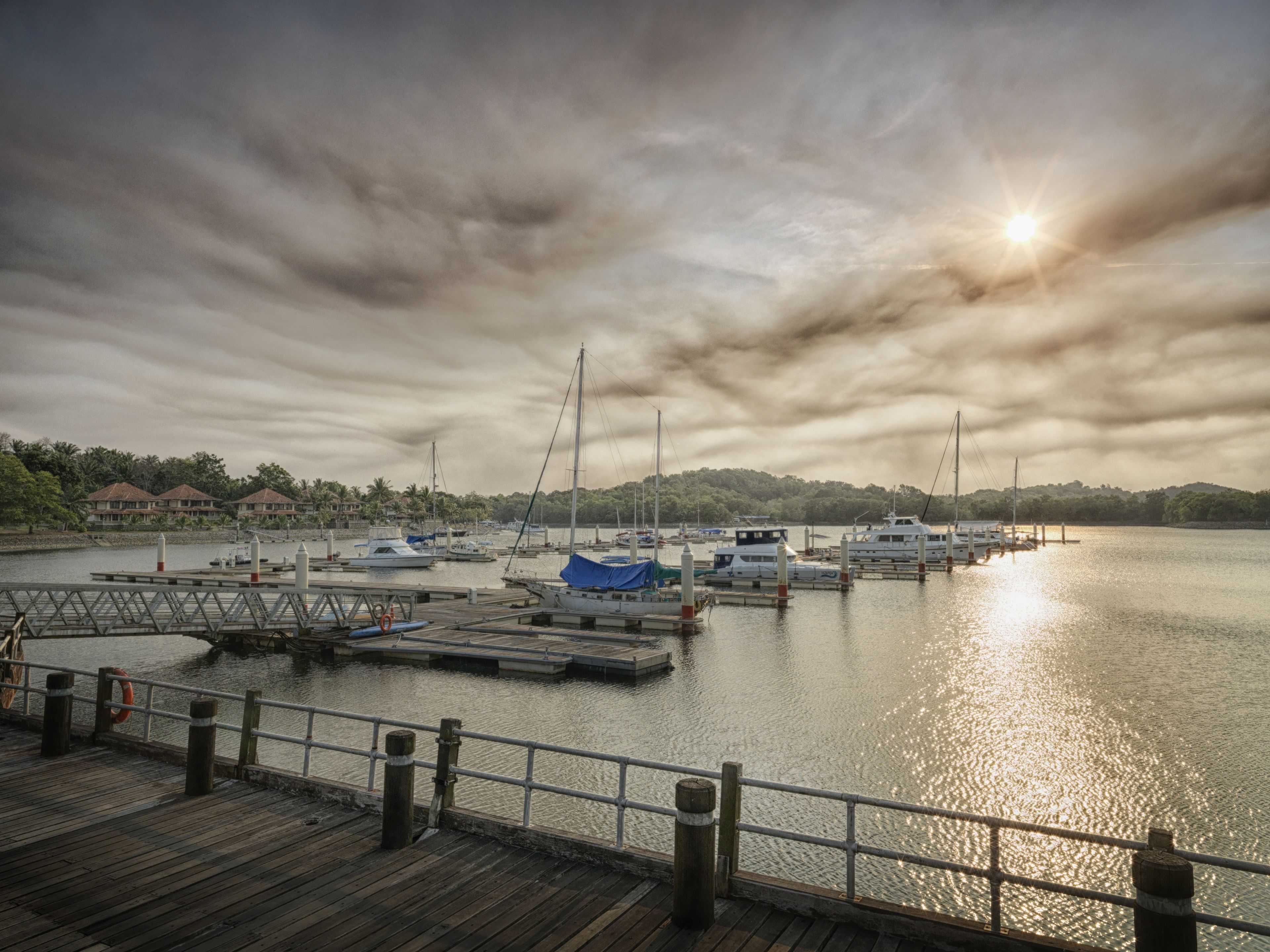 A marina with boats docked, a wooden pier, and a cloudy sky with the sun visible.