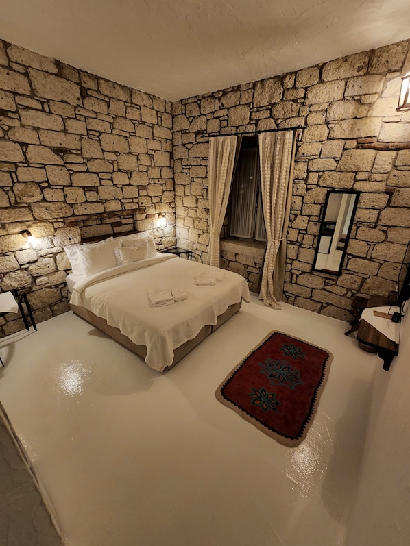 Une chambre avec un mur en pierre, un lit recouvert de draps blancs, un tapis rouge et un miroir au mur.