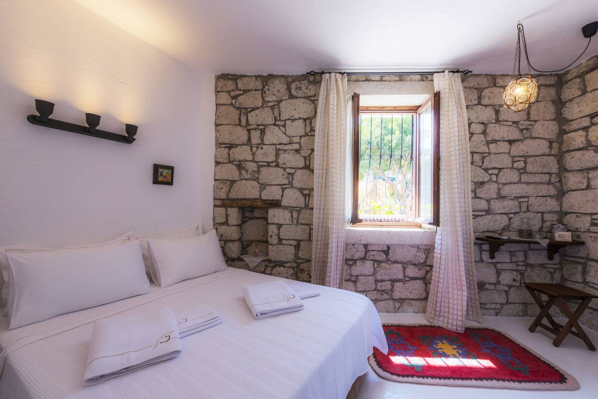 Una habitación con pared de piedra, cama con ropa de cama blanca, ventana con cortinas y una lámpara colgante.