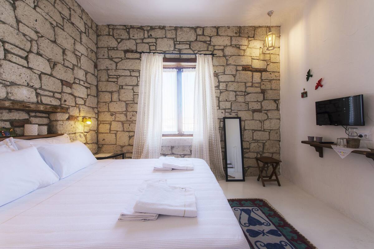 Un dormitorio con pared de piedra, una cama con ropa blanca, una mesita de noche con lámpara, un televisor en la pared y una ventana con cortinas.