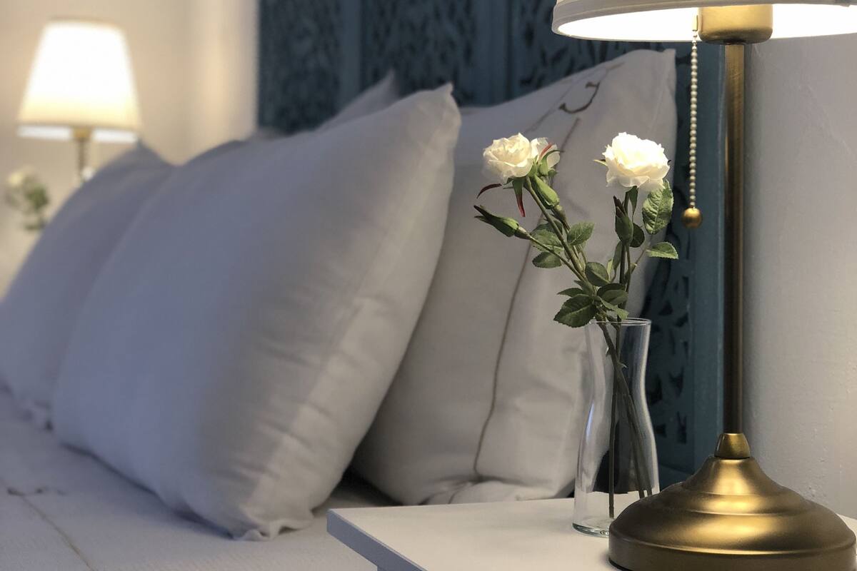 Una cama bien hecha con sábanas blancas, una mesita de noche con una lámpara y un jarrón con flores blancas.