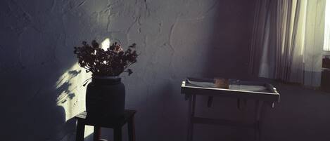 A rustic room with a wooden stool, a vase with dried flowers, and a table with a glass and a container.