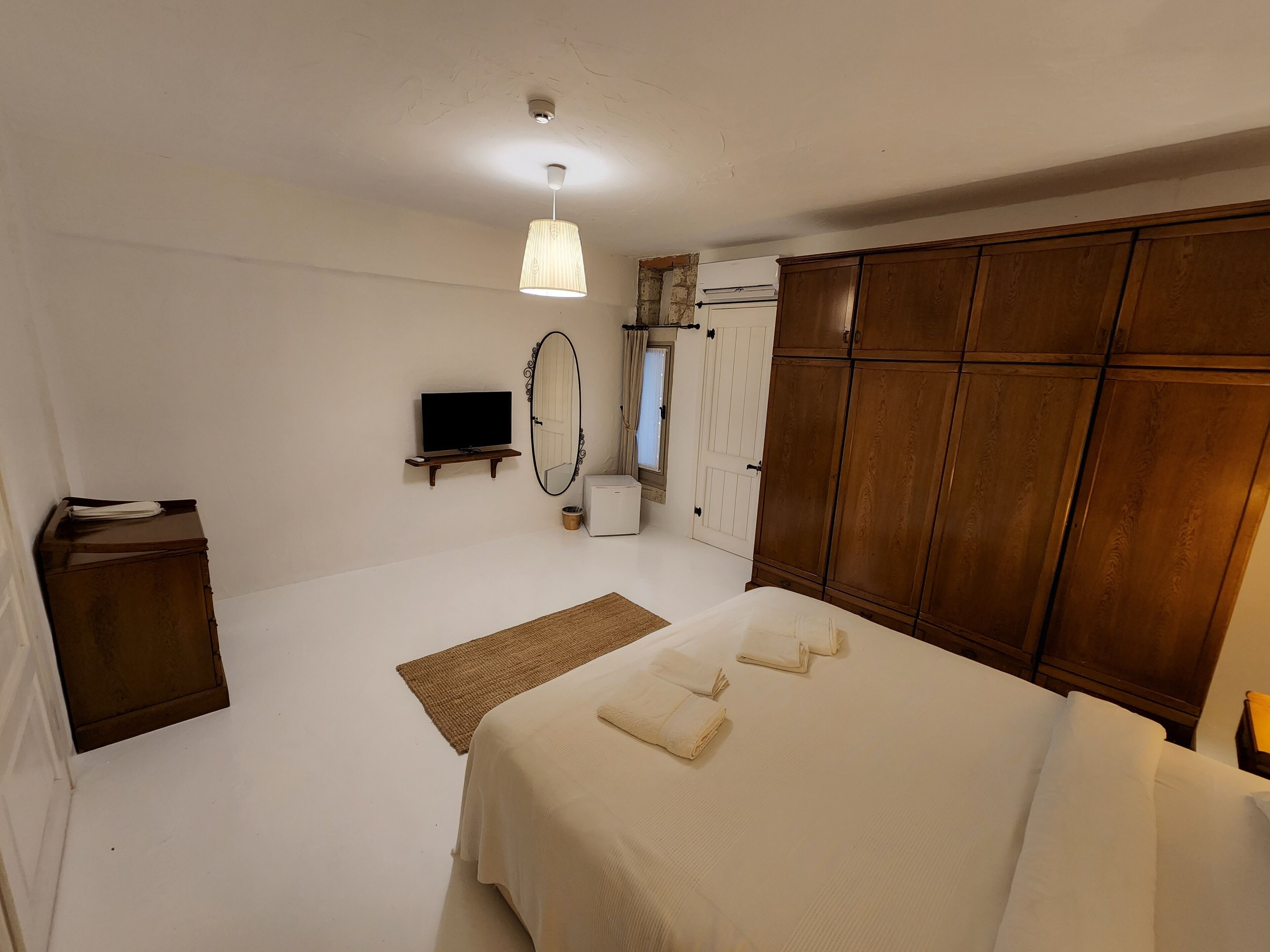 A bedroom with a bed, a wooden wardrobe, a television on a stand, a mirror, and a small heater.