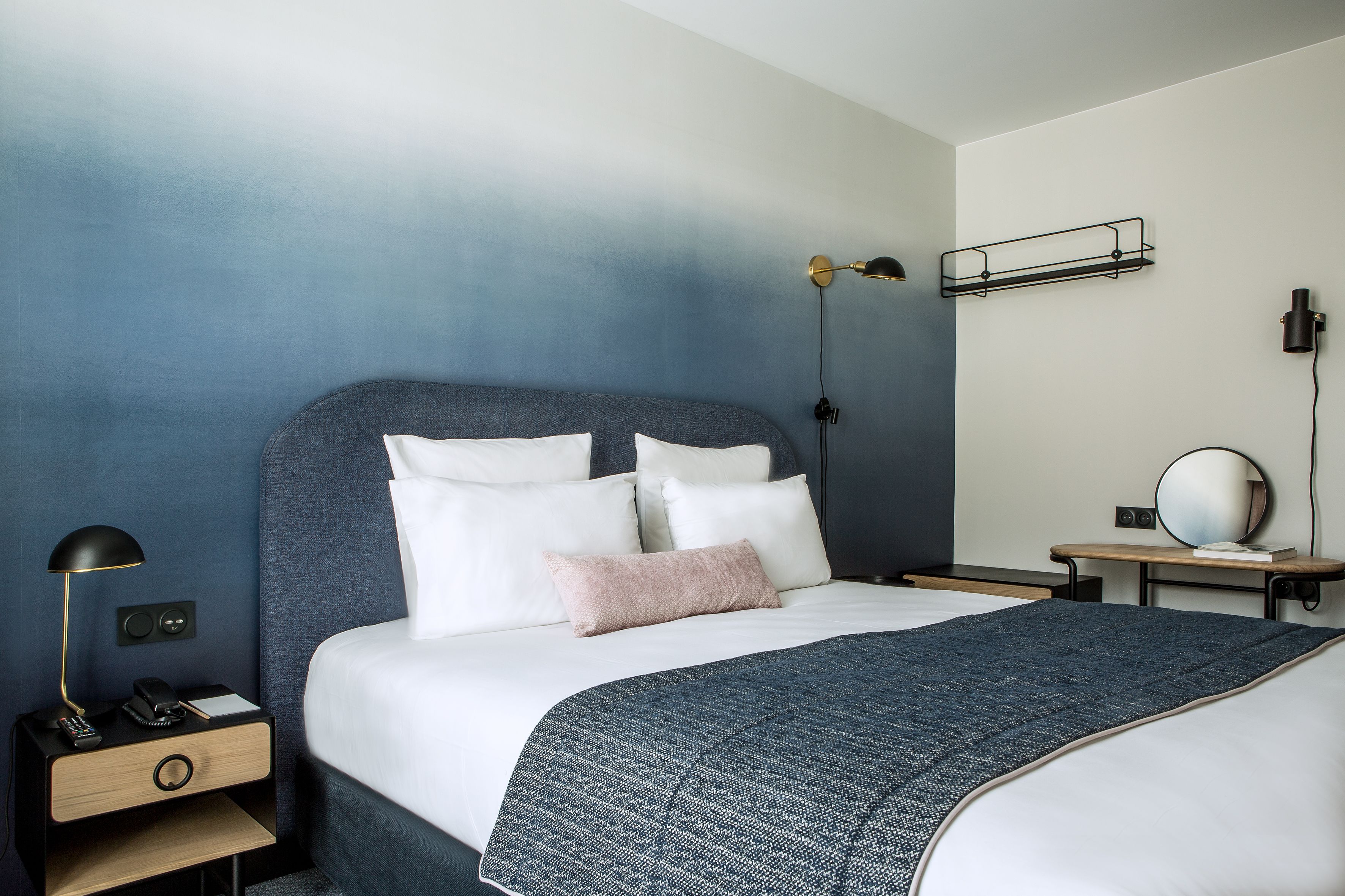 A neatly made bed with a blue headboard, a bedside table with a lamp, and a wall-mounted shelf with a mirror.