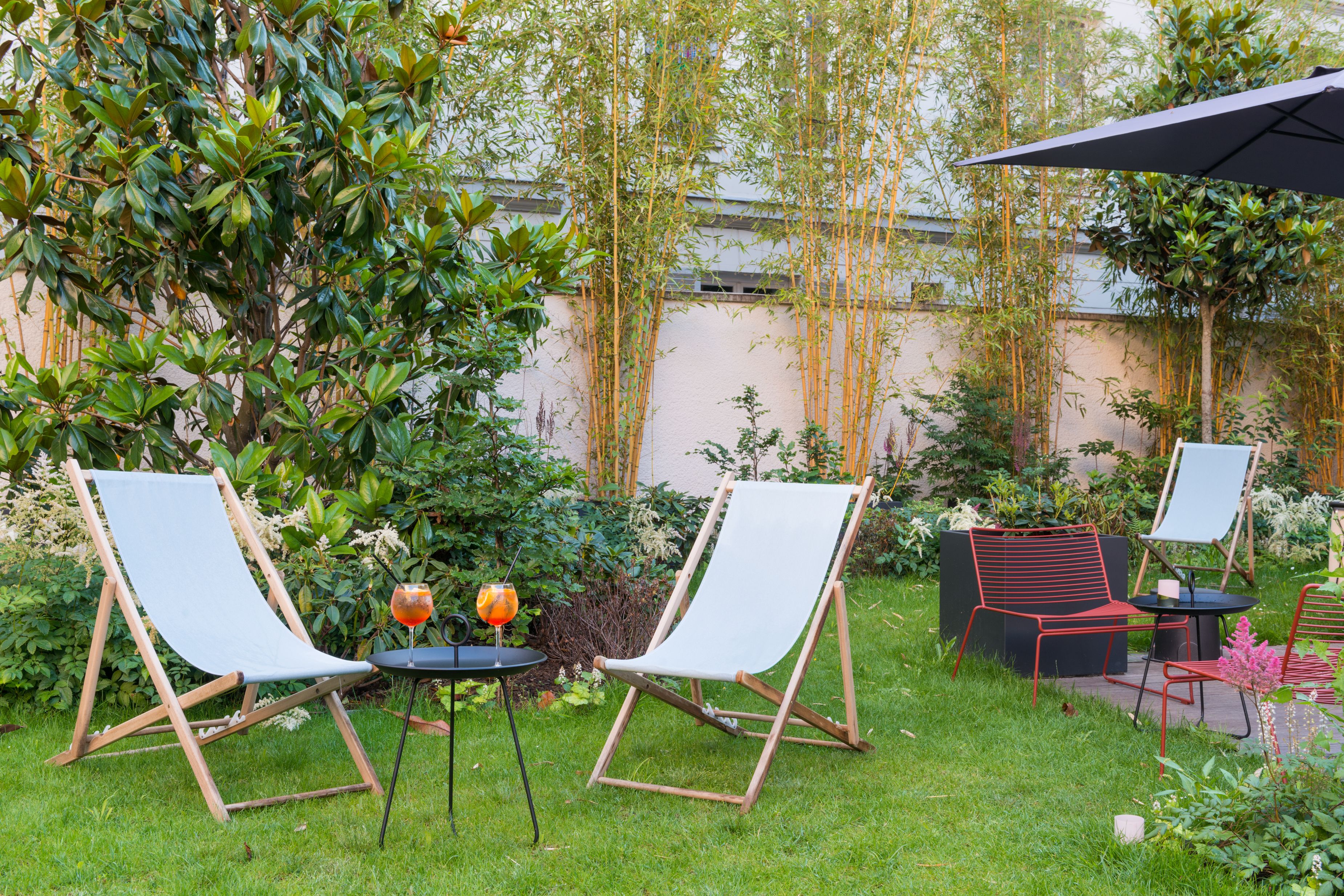 A garden with two white canvas chairs, a small table with a drink, and a red metal chair.