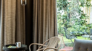 Quarto moderno com vista para o verde, com uma cadeira de vime, uma poltrona cinza e uma mesinha com uma xícara e um copo amarelo.