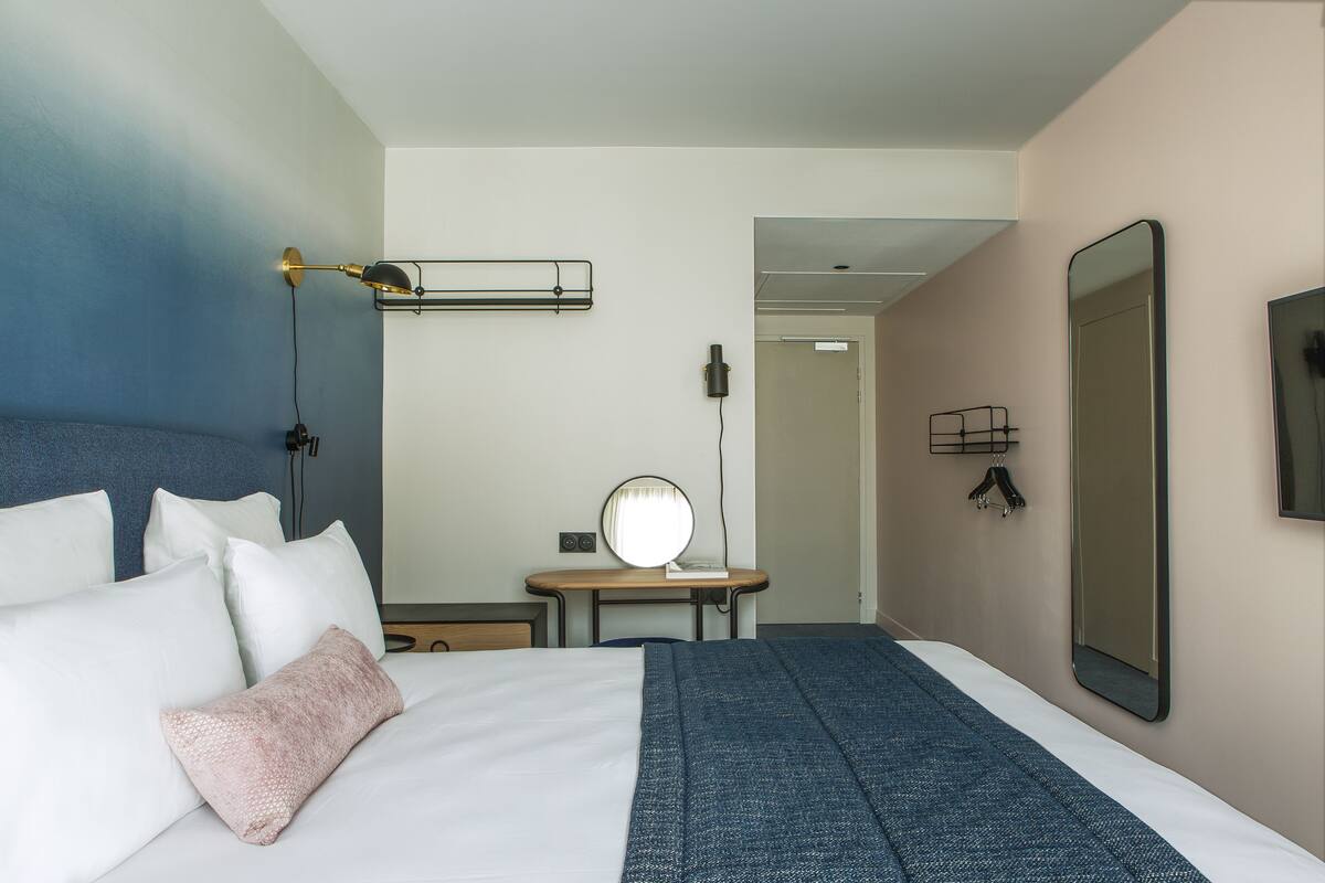 A hotel room with a bed, a bedside table, a mirror, and a wall-mounted TV.