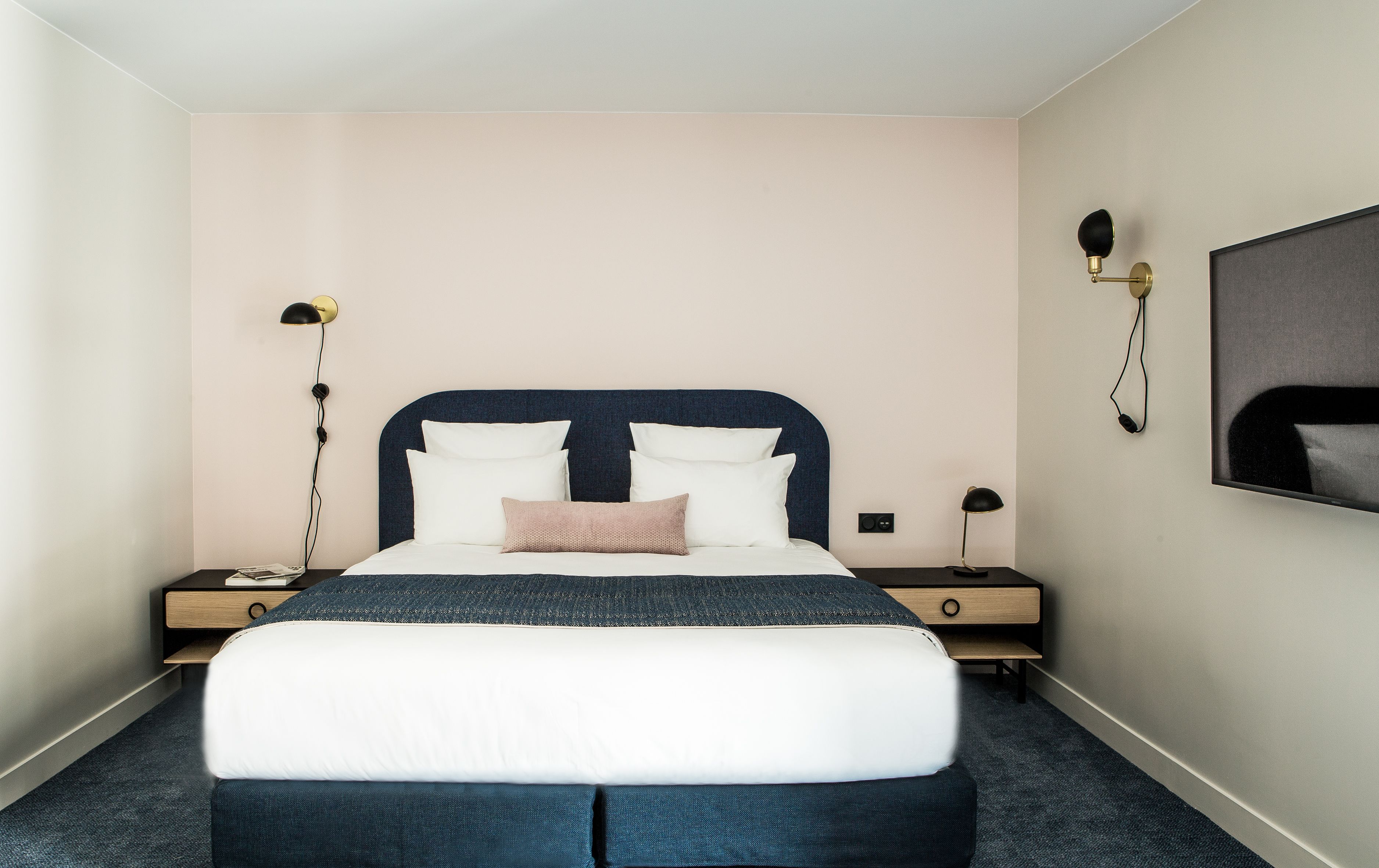 Habitación de hotel ordenada con una cama grande, mesitas de noche, un televisor montado en la pared y una lámpara.