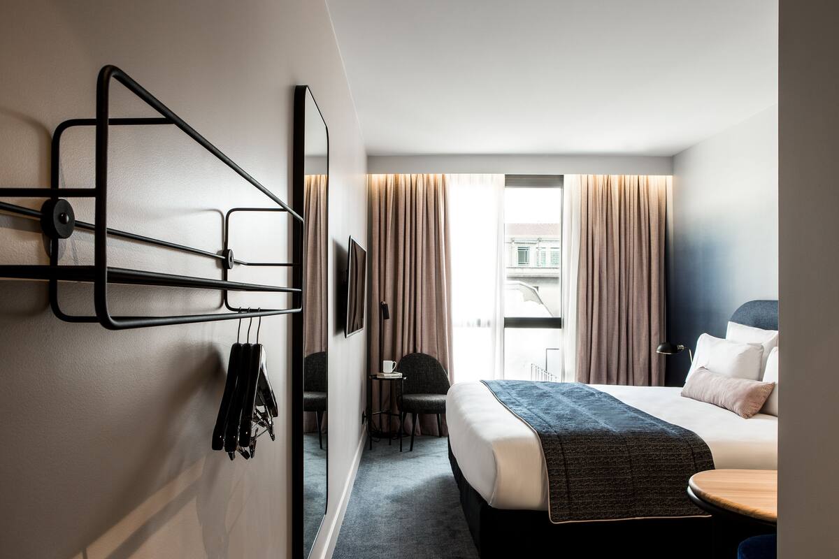 A hotel room with a bed, a desk, a chair, a TV mounted on the wall, and a window with curtains.