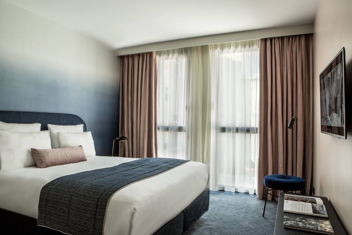 A neatly made bed with a dark headboard, white pillows, and a brown throw blanket. There is a small stool and a framed picture on the wall.