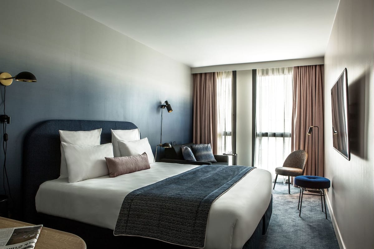 A hotel room with a large bed, a desk, a chair, a TV, and a window with curtains.