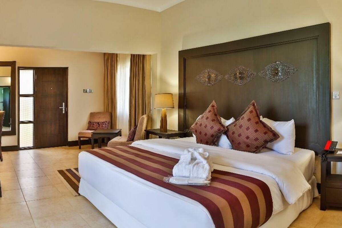 A hotel room with a large bed, a desk, a chair, and a nightstand with a lamp.