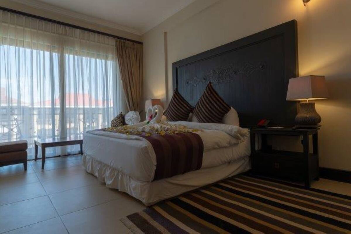 A hotel room with a large bed, a bedside table with a lamp, a window with curtains, and a view of the cityscape.