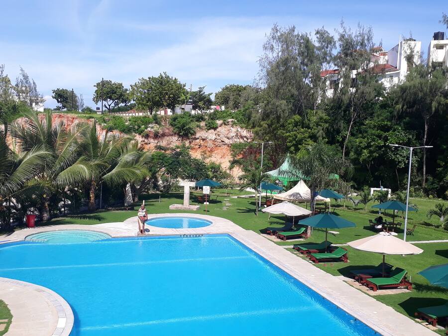 Ein Swimmingpool mit Liegestühlen und Sonnenschirmen, umgeben von Grünflächen und Gebäuden im Hintergrund.