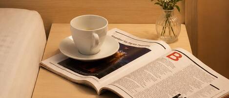 Una cama bien tendida, una mesita de noche con una taza de café, un jarrón con flores y una revista.