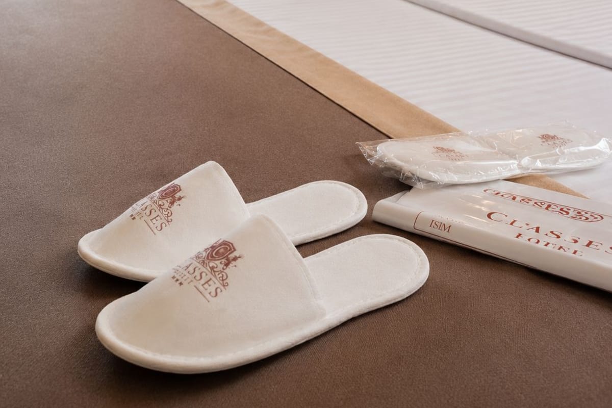 A hotel room with a bed, a chair, and slippers on the bed.