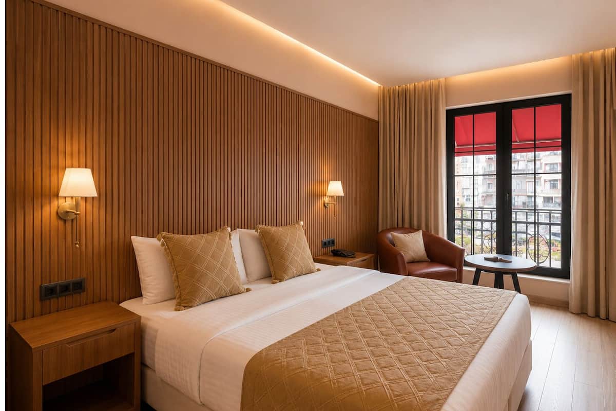 A hotel room with a large bed, two bedside lamps, a small table, a chair, and a window with a view of buildings.