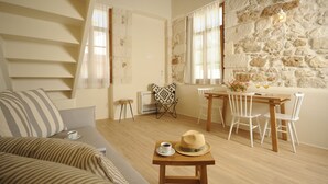 A cozy living room with a sofa, a dining table with chairs, a small table with a cup and a hat, and a stone wall. - Veranda Rossa Suites (Rethymno)