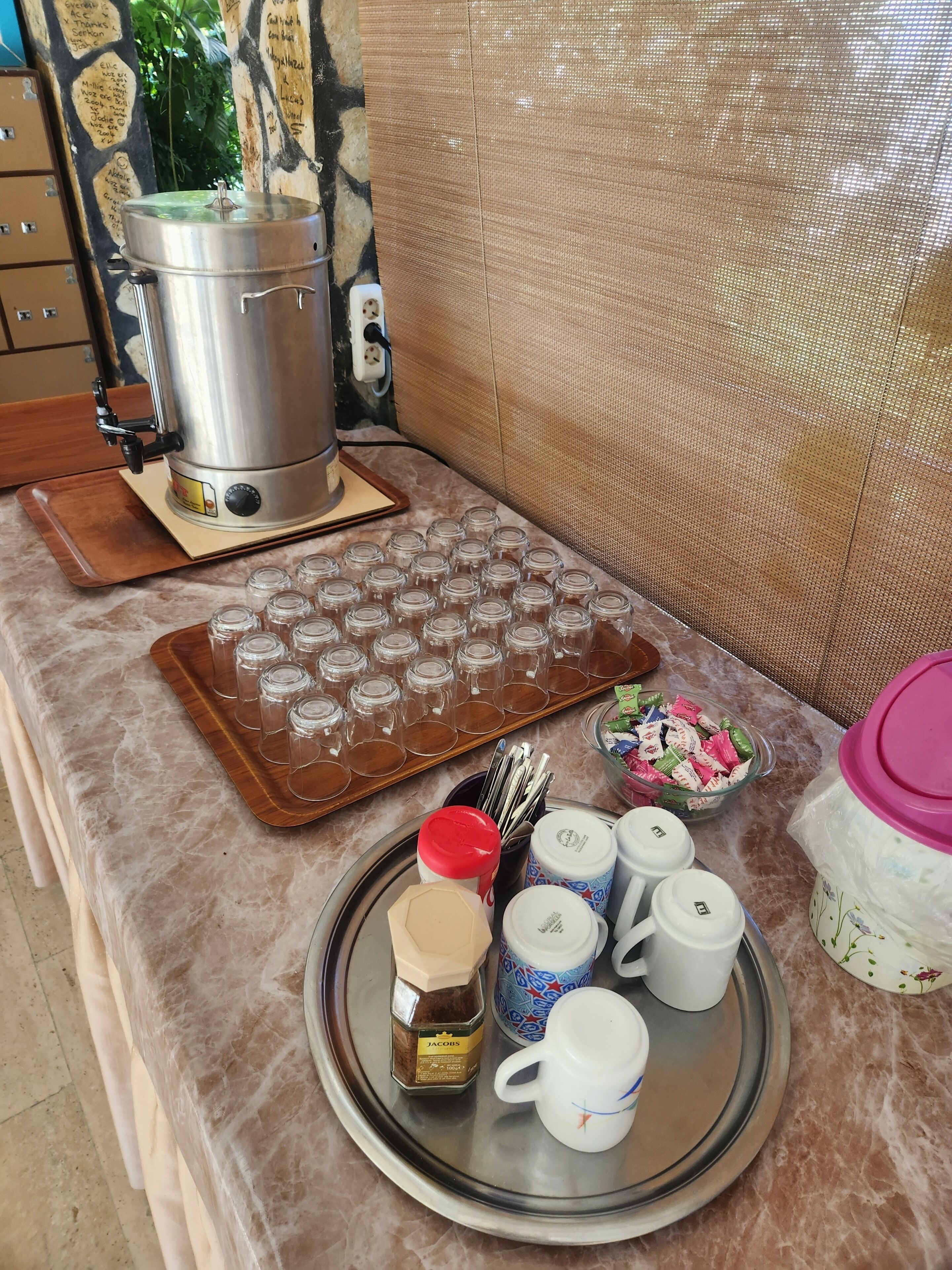 A kitchen counter with a coffee maker, a tray of empty glasses, a jar of coffee, a mug, and a bowl of snacks.