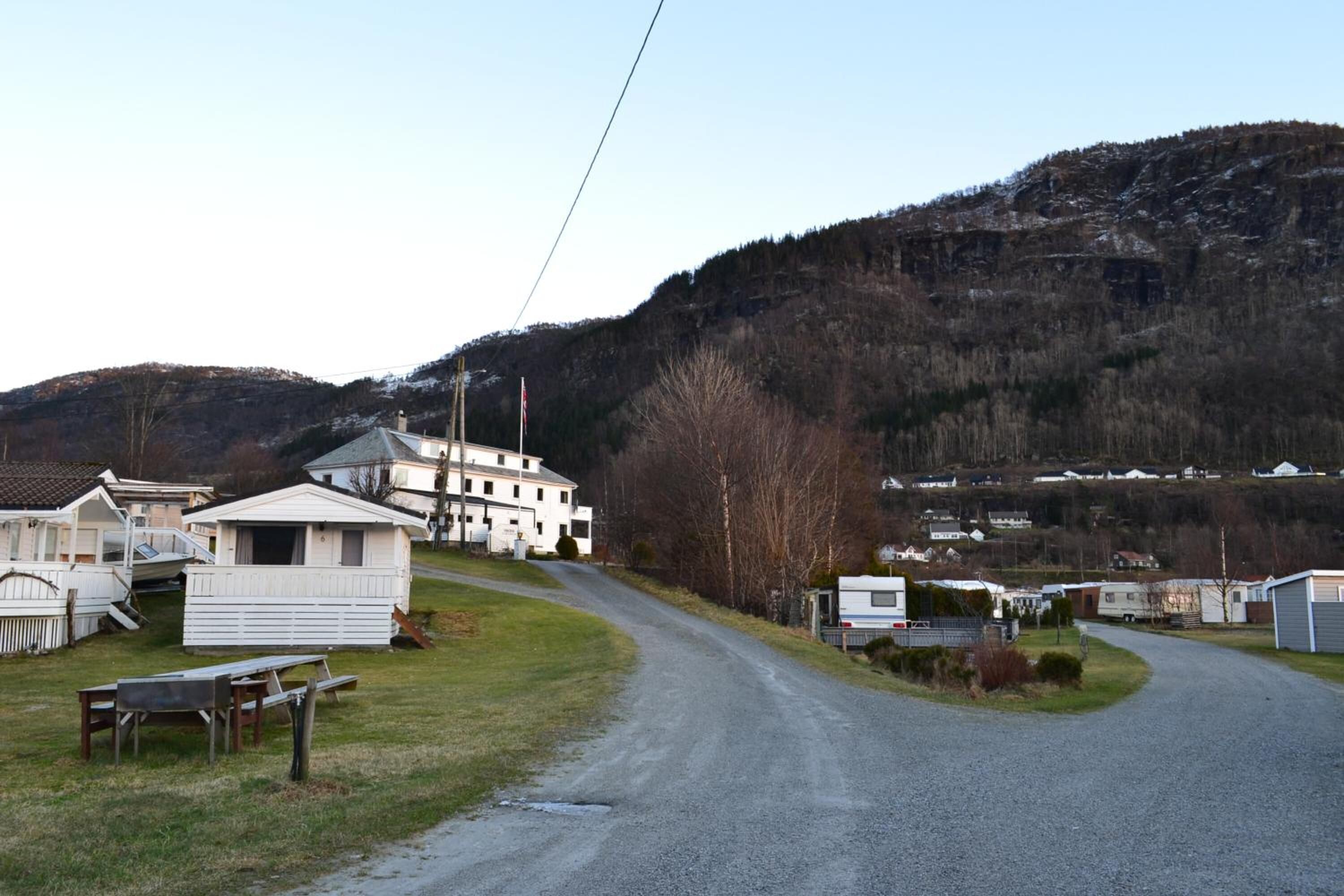 A gravel road leading to a cluster of white mobile homes with a large building in the background.