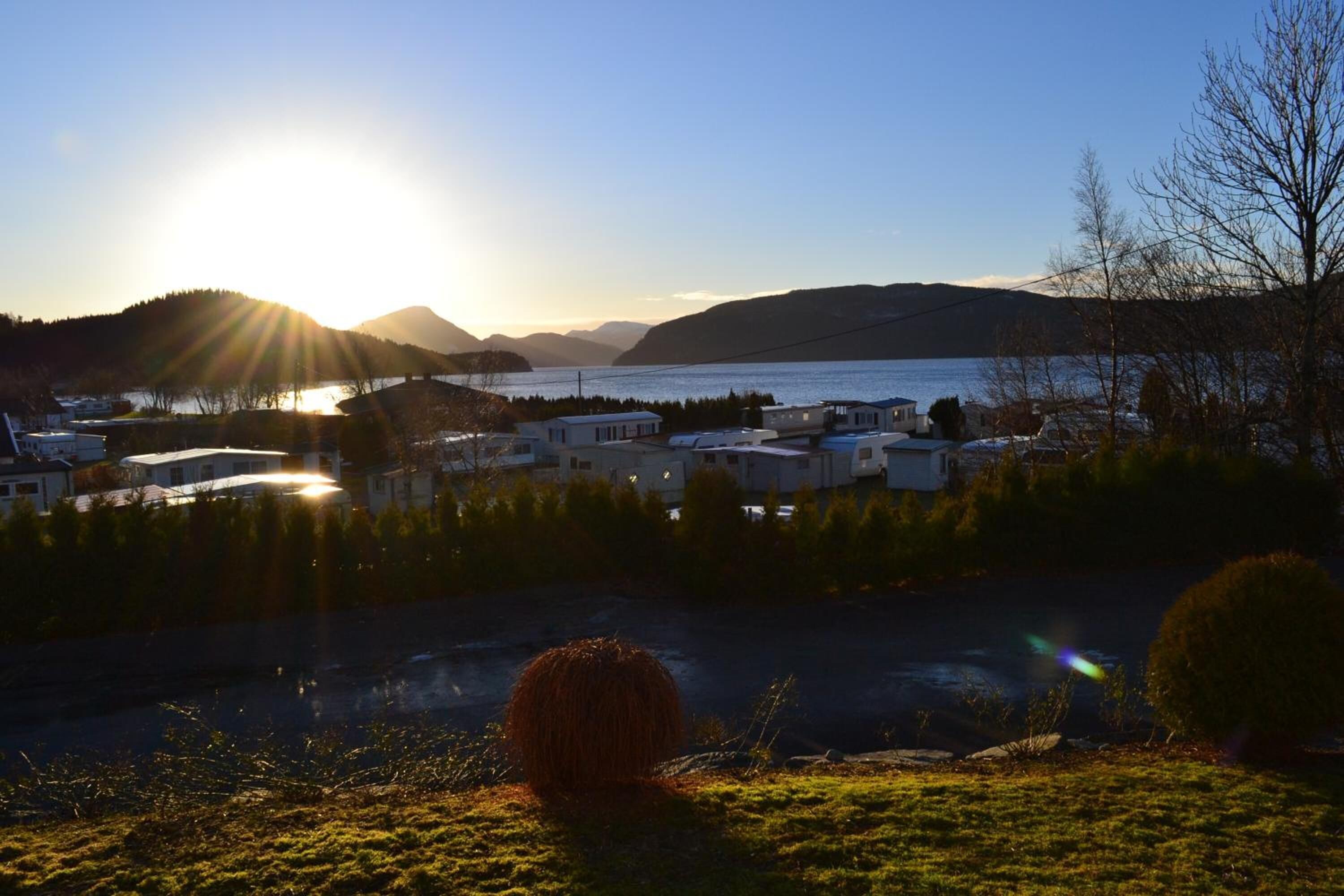 A lakeside view with a clear sky, a sun setting or rising over the water, and a small cluster of buildings in the foreground.