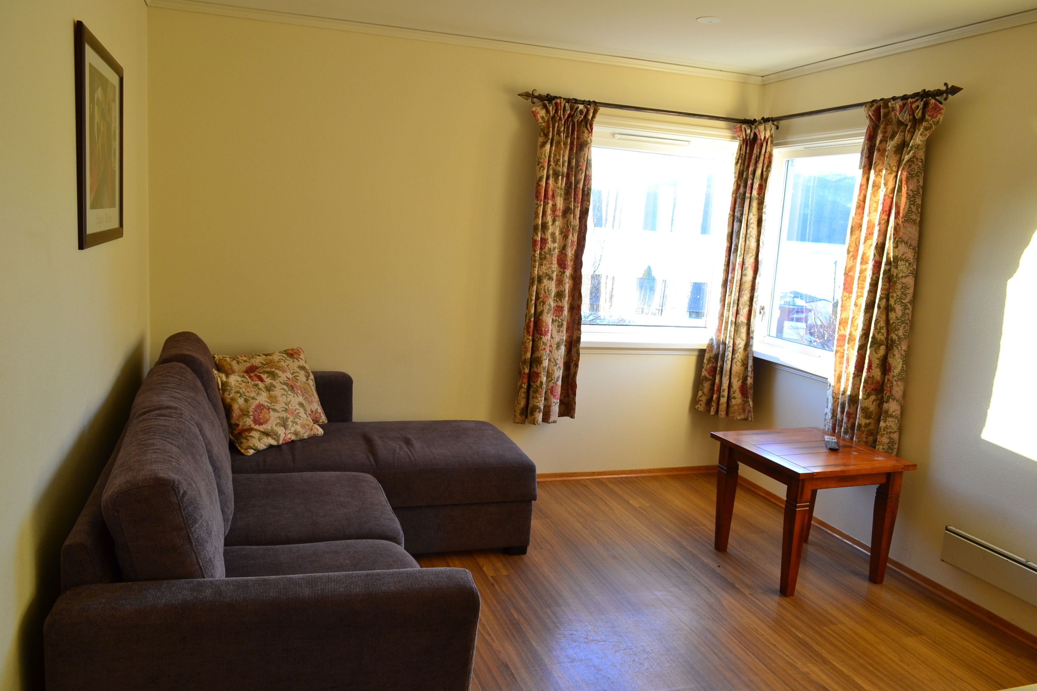 A living room with a brown sectional sofa, a small wooden table, and a window with floral curtains.