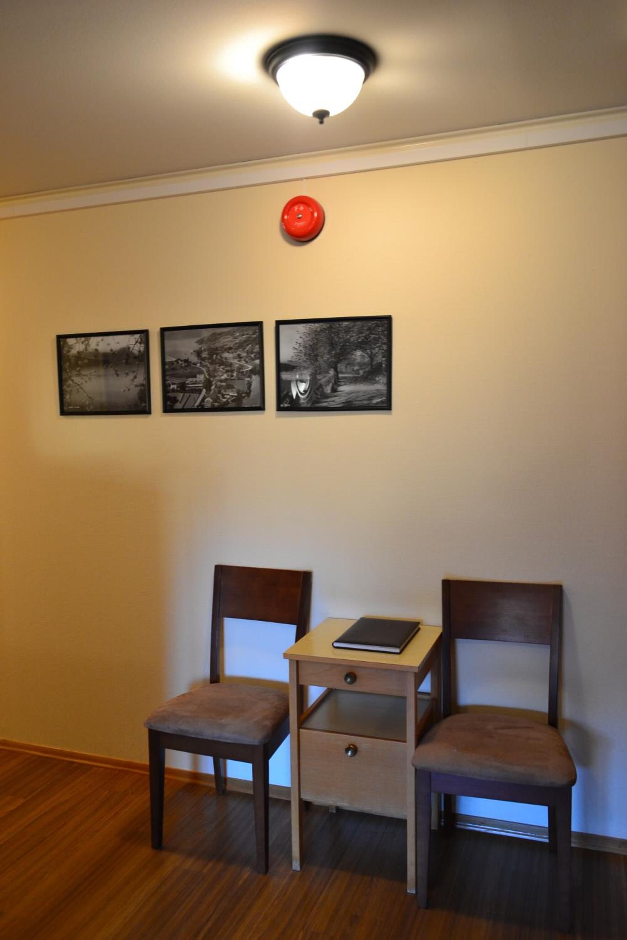 A room with a wooden floor, two chairs, a small table with a book, and three black-and-white framed pictures on the wall.