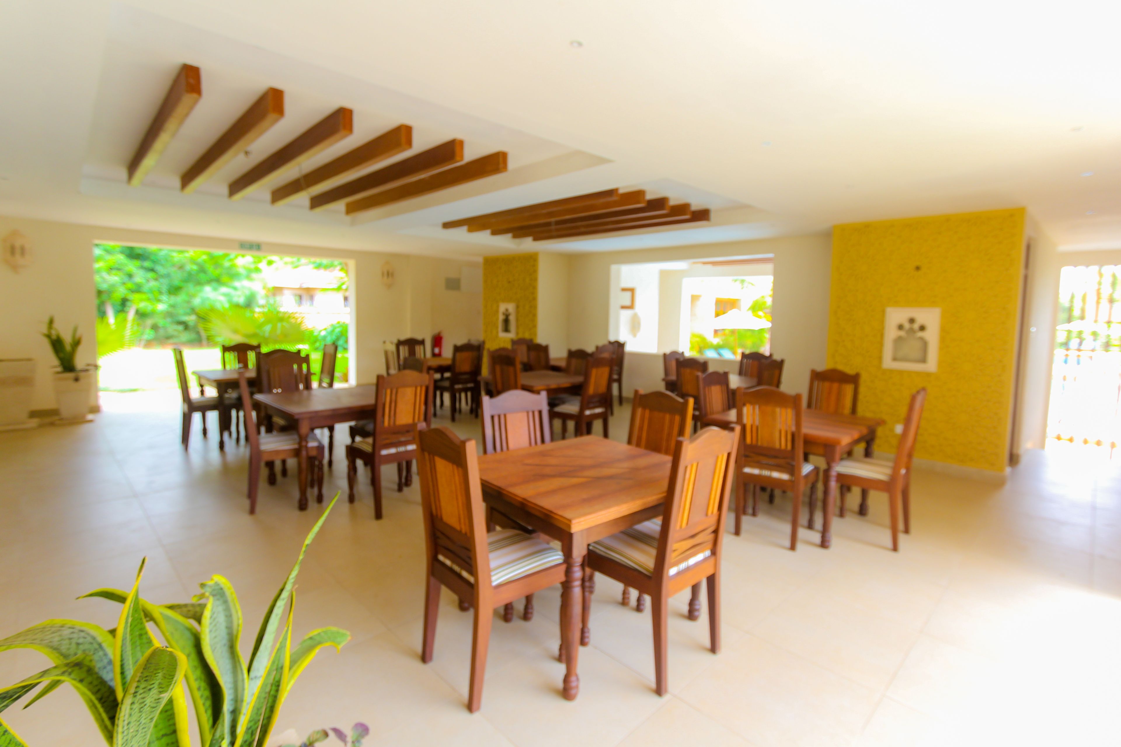 A spacious dining area with multiple wooden tables and chairs, a yellow accent wall, and large windows allowing natural light.