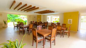 A spacious dining area with multiple wooden tables and chairs, a yellow accent wall, and large windows allowing natural light.
