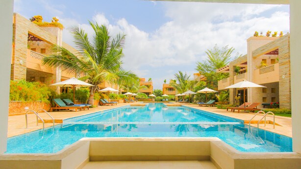 Una piscina rodeada de sillas de descanso y sombrillas, con palmeras y edificios al fondo.