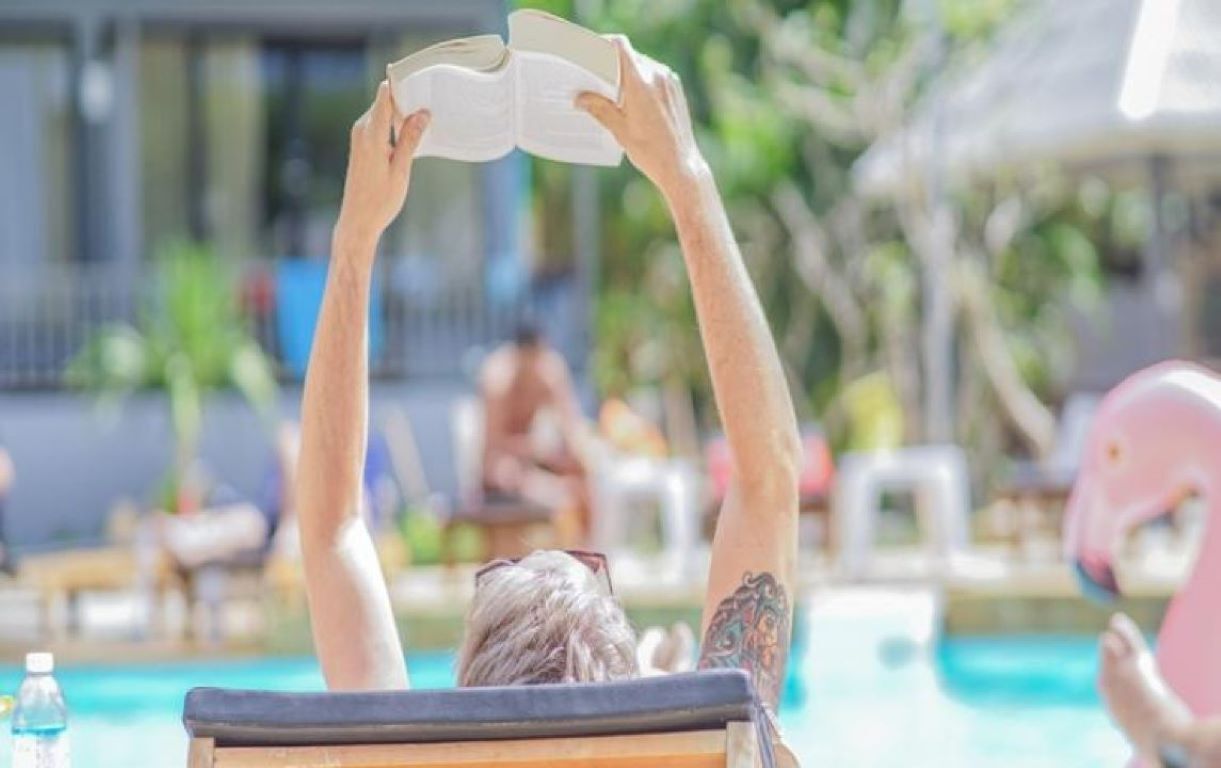 Une personne est allongée au bord d’une piscine, un livre à la main, et elle a devant elle un espace piscine aménagé avec des chaises longues et des parasols.