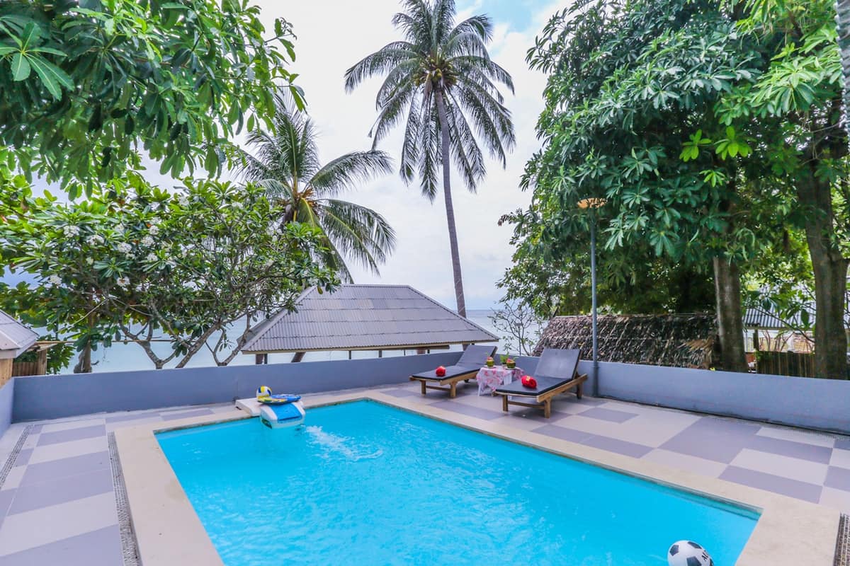 A swimming pool with lounge chairs and a table, surrounded by palm trees and a view of the ocean.