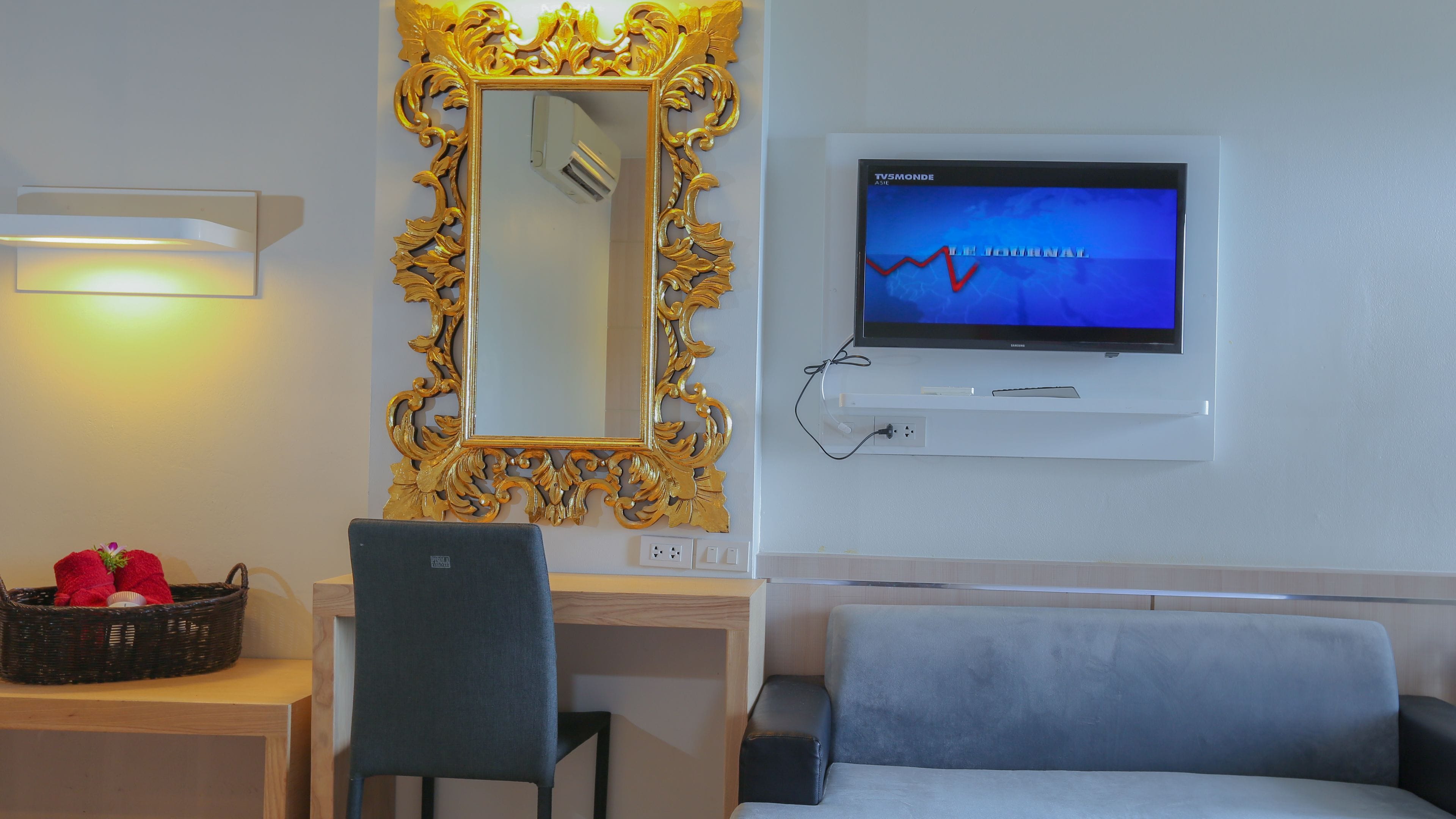 Habitación de hotel con televisor montado en la pared, escritorio con silla, sofá y espejo decorativo con marco ornamentado.