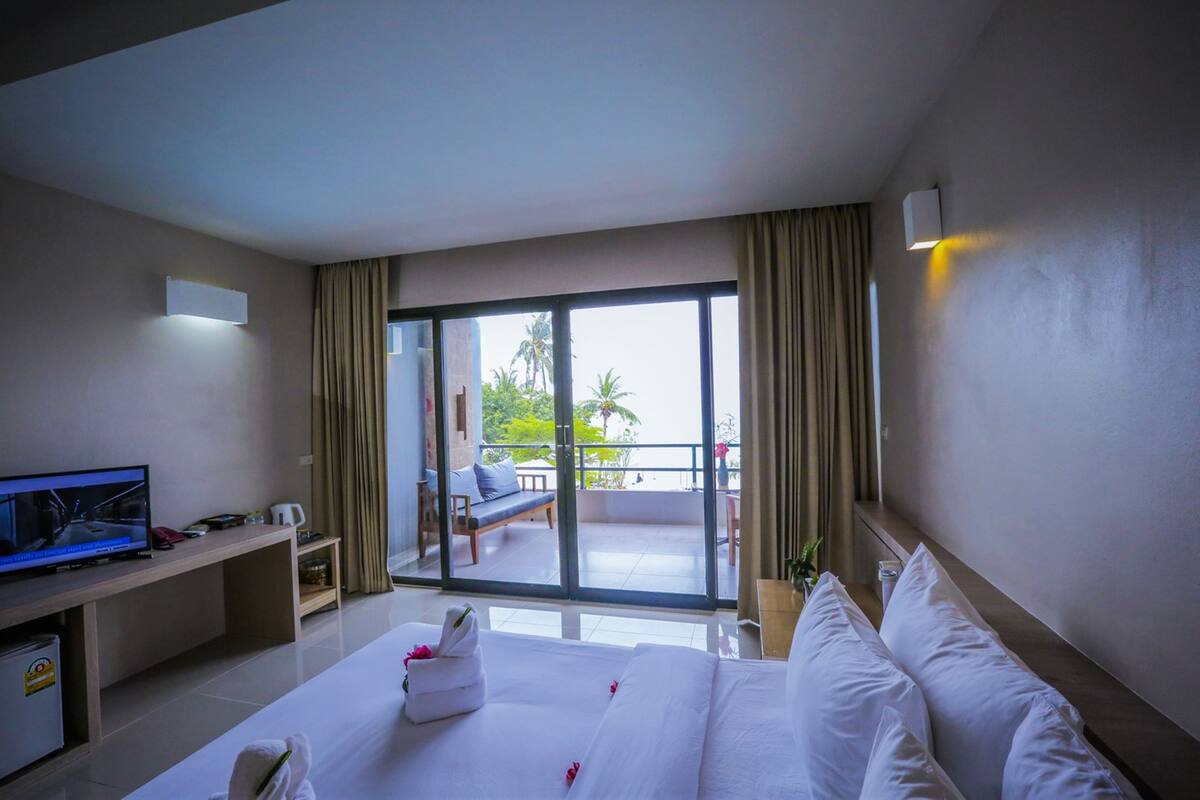 A hotel room with a bed, a television, a desk, a balcony with a chair and a table, and a view of palm trees.
