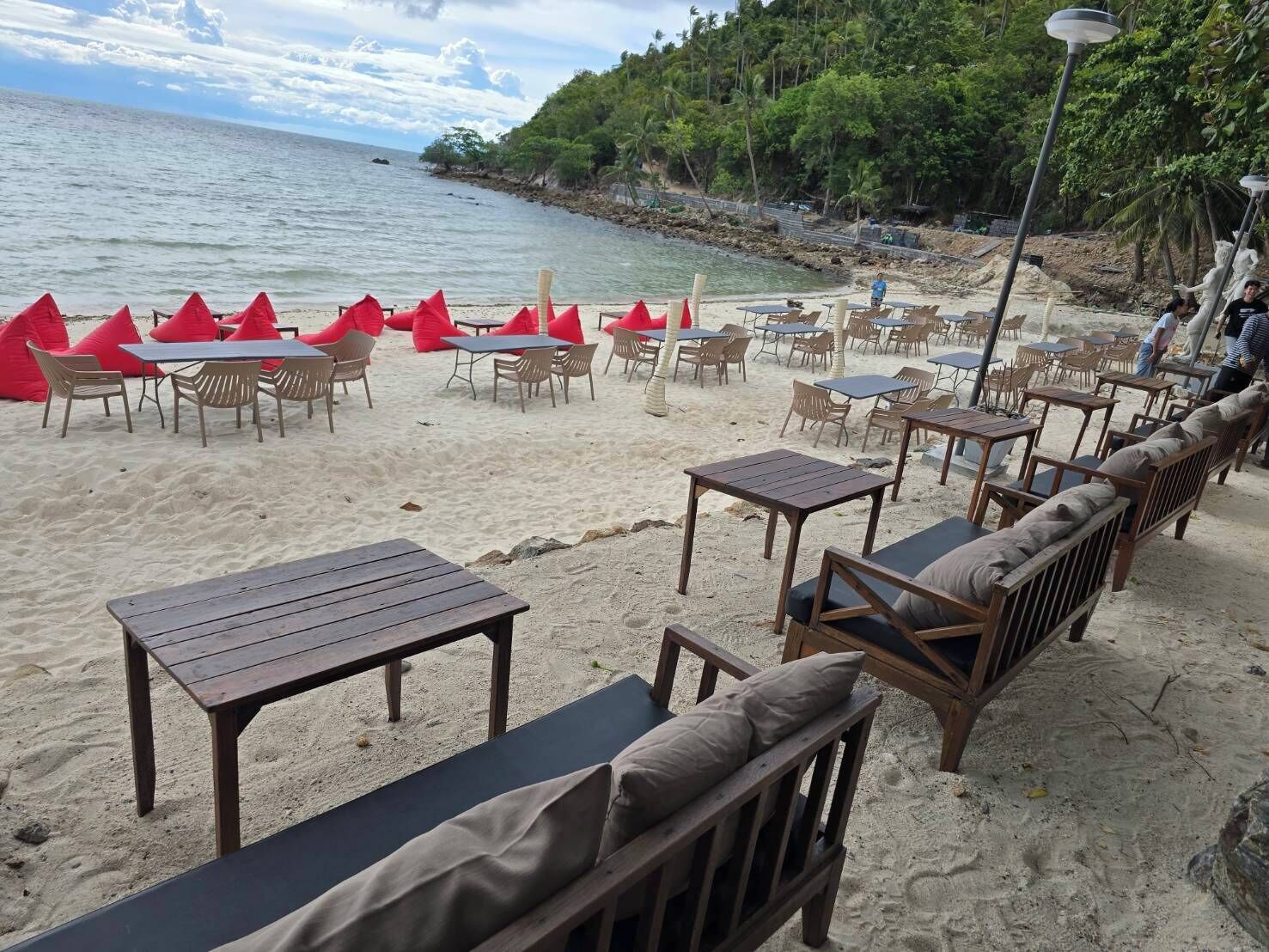A beachside café with wooden tables and chairs, a sandy area, and a view of the sea.