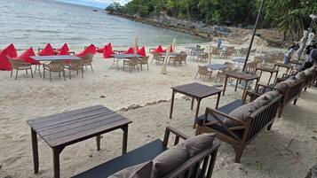 A beachside café with wooden tables and chairs, a sandy area, and a view of the sea.