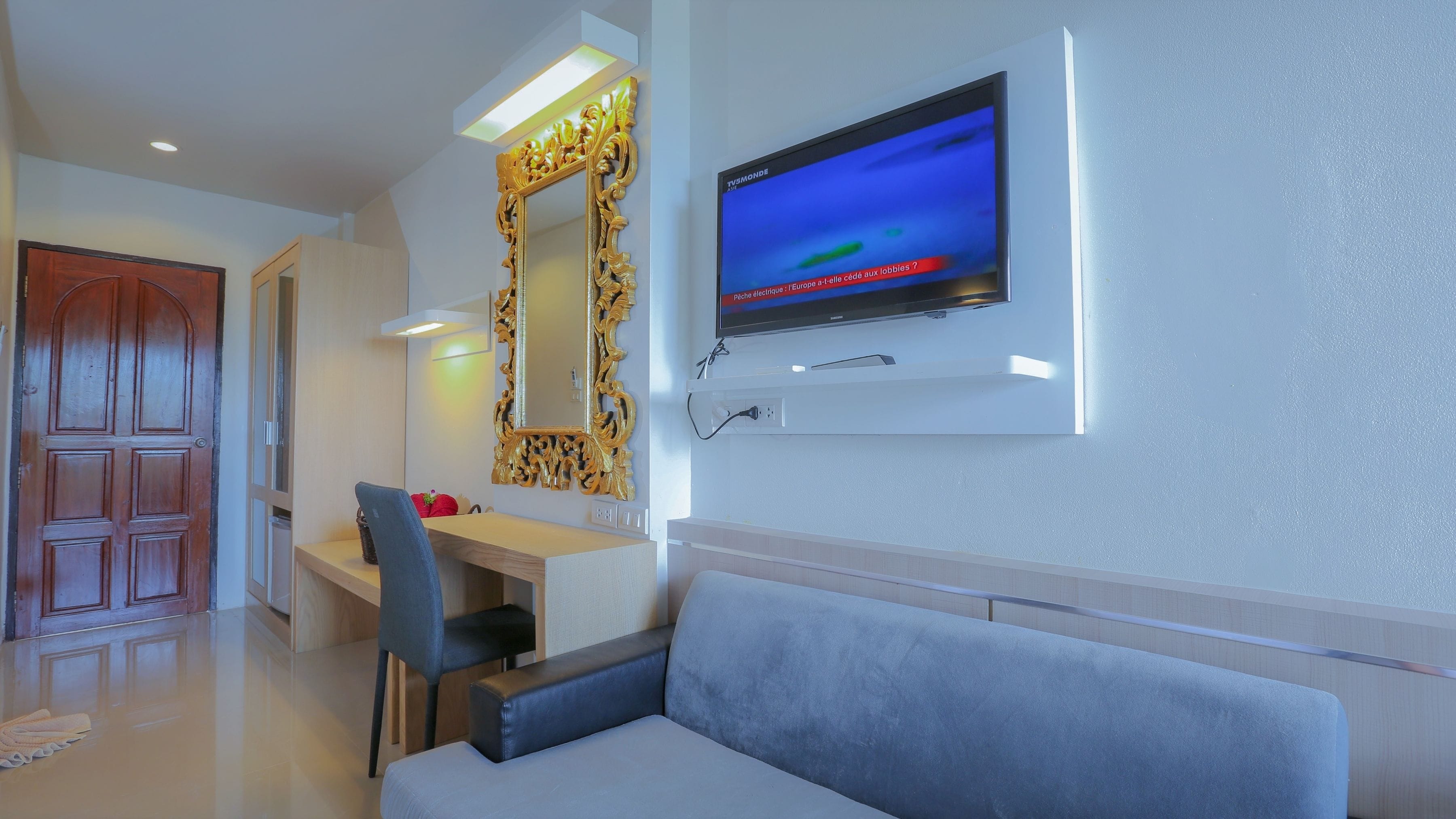 A modern hotel room with a flat-screen TV mounted on the wall, a wooden desk, a blue chair, and a grey sofa.