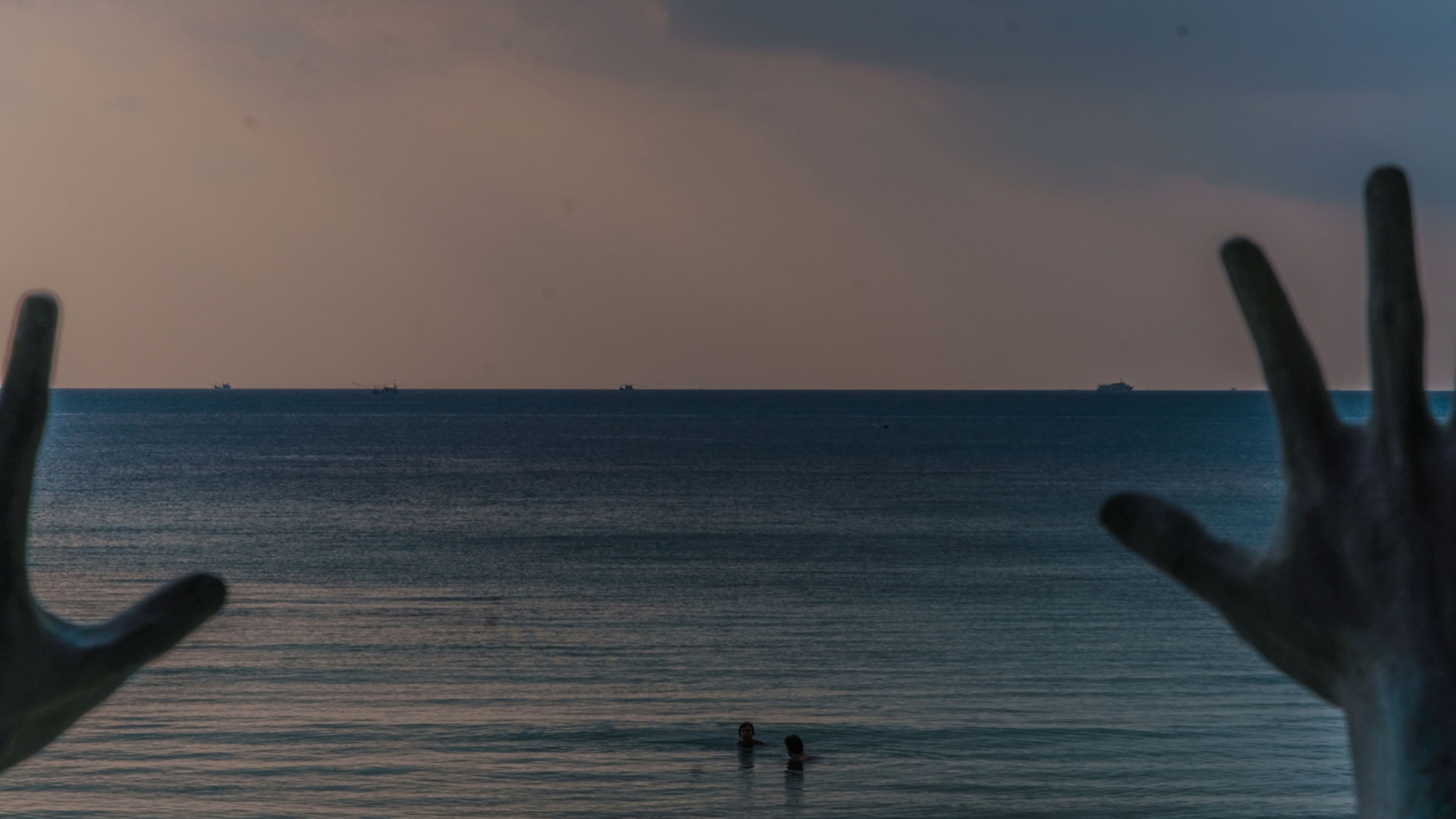 Une vue sur la mer, avec deux personnes dans l’eau et des navires à l’horizon.