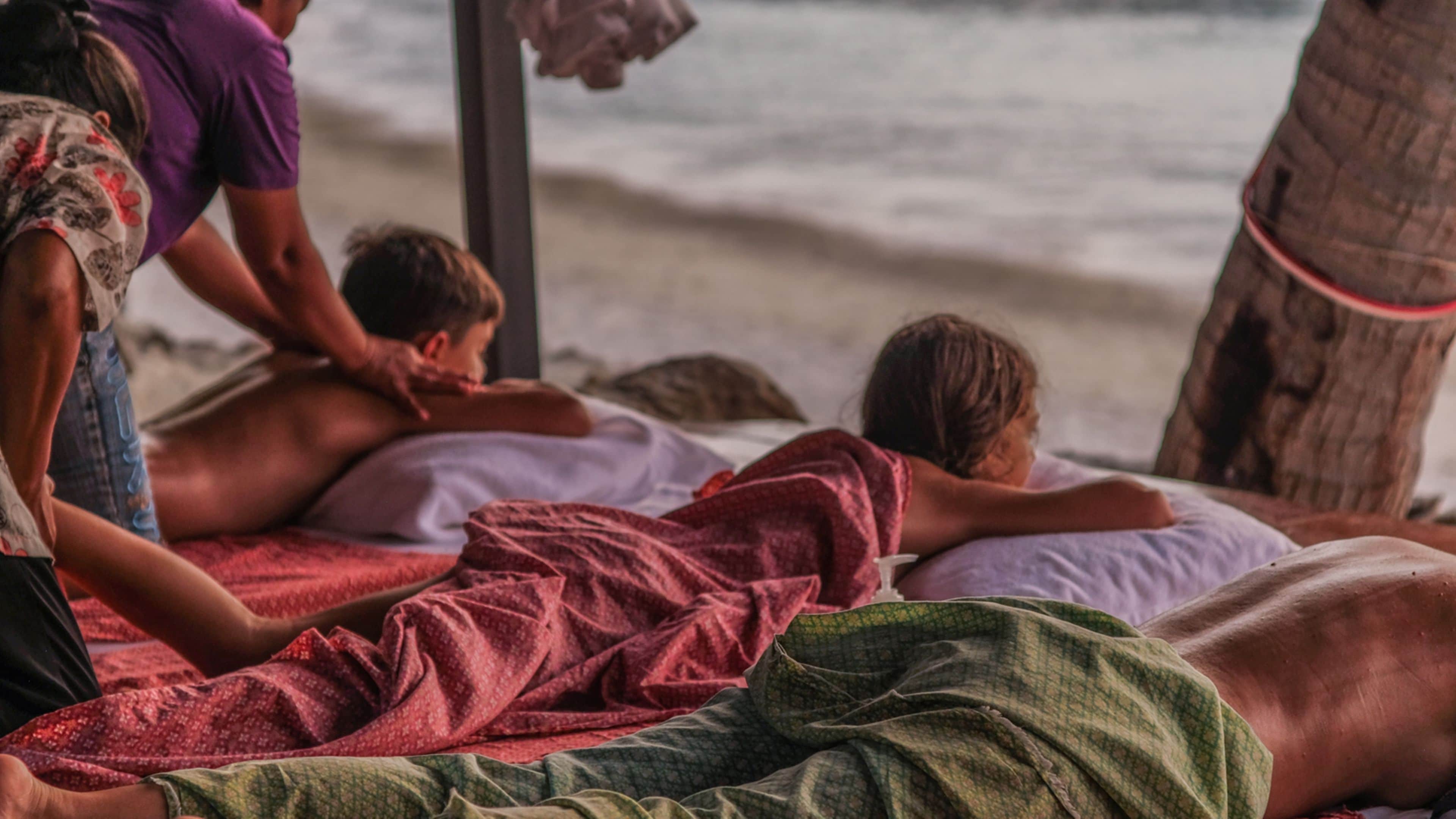 A beachside massage setup with individuals lying on mats, covered with blankets, and a wooden structure in the background.