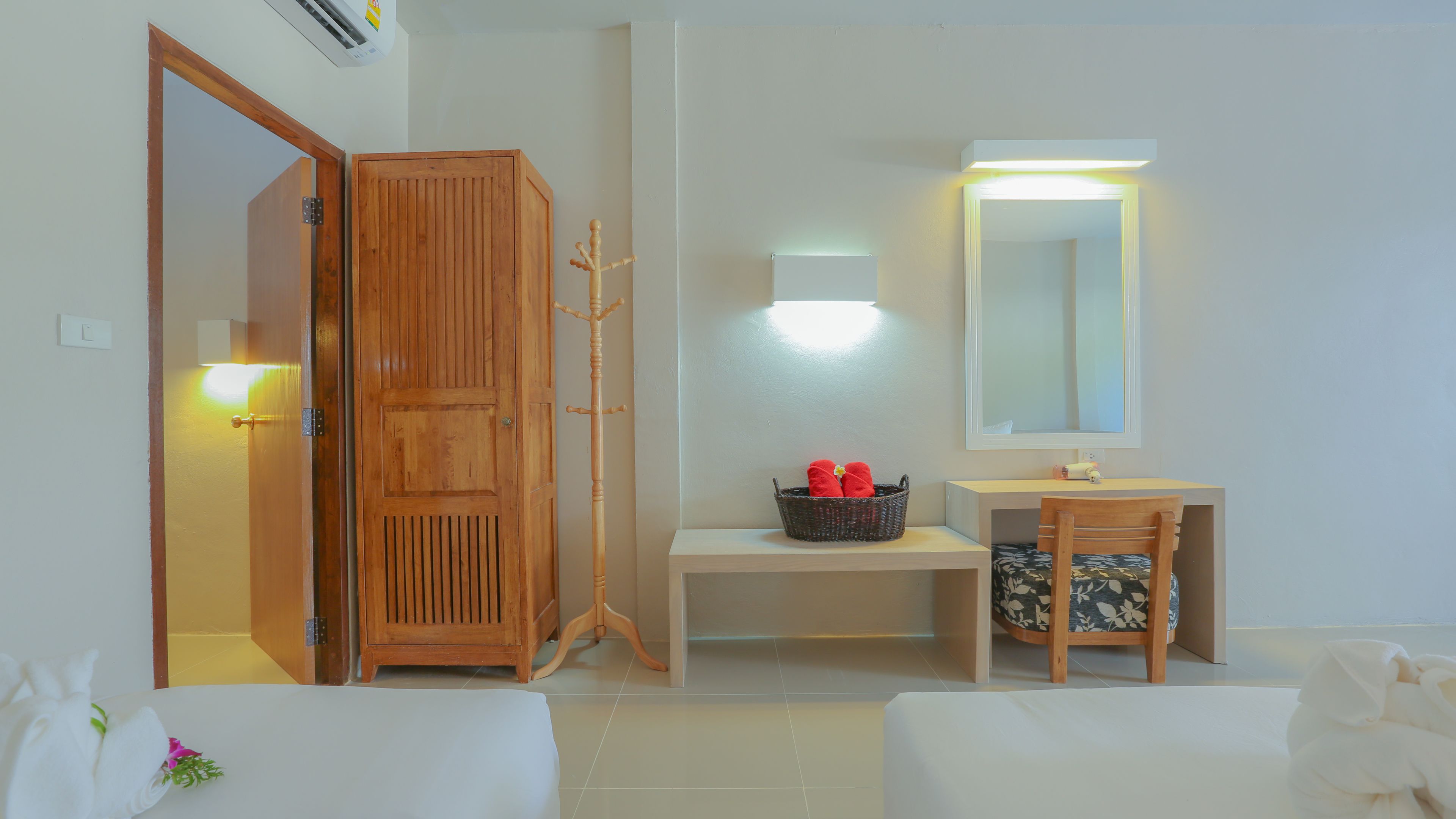 A room with a wooden wardrobe, a desk with a basket, a chair, a mirror, and a bed.