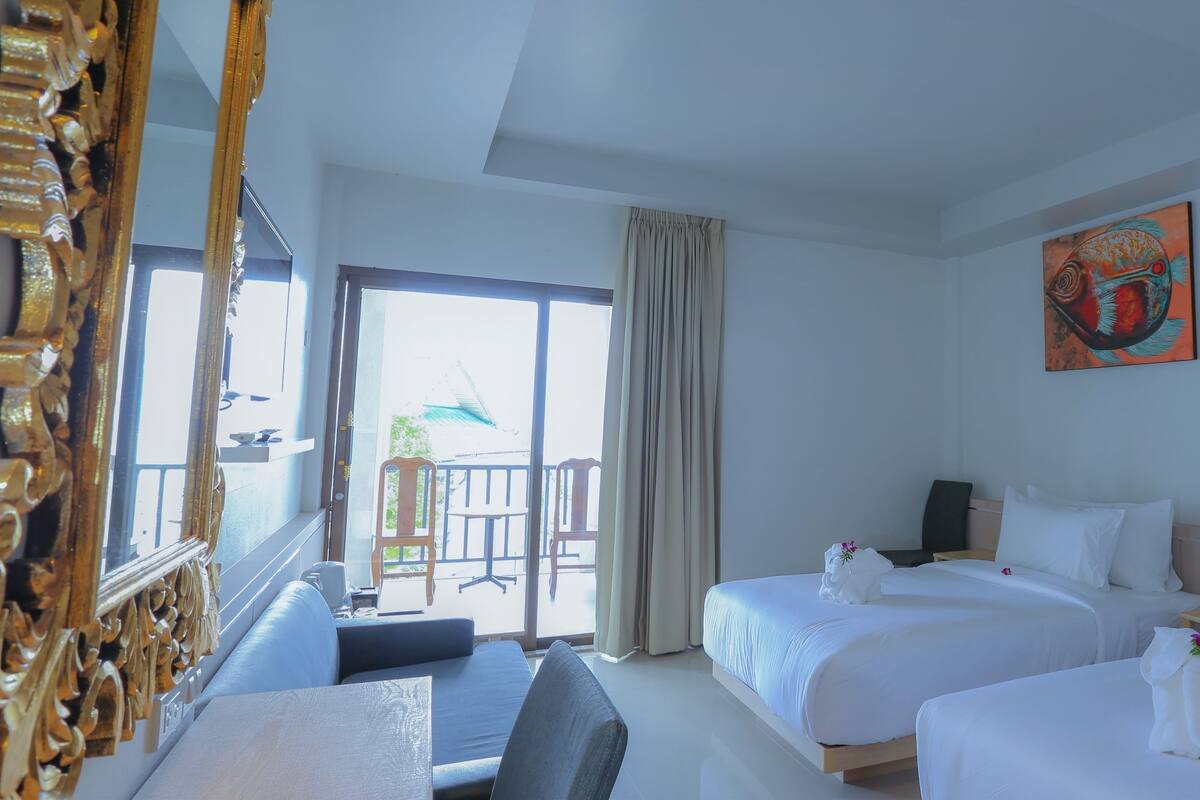 A hotel room with two beds, a desk, a chair, a balcony with a view, and a painting on the wall.