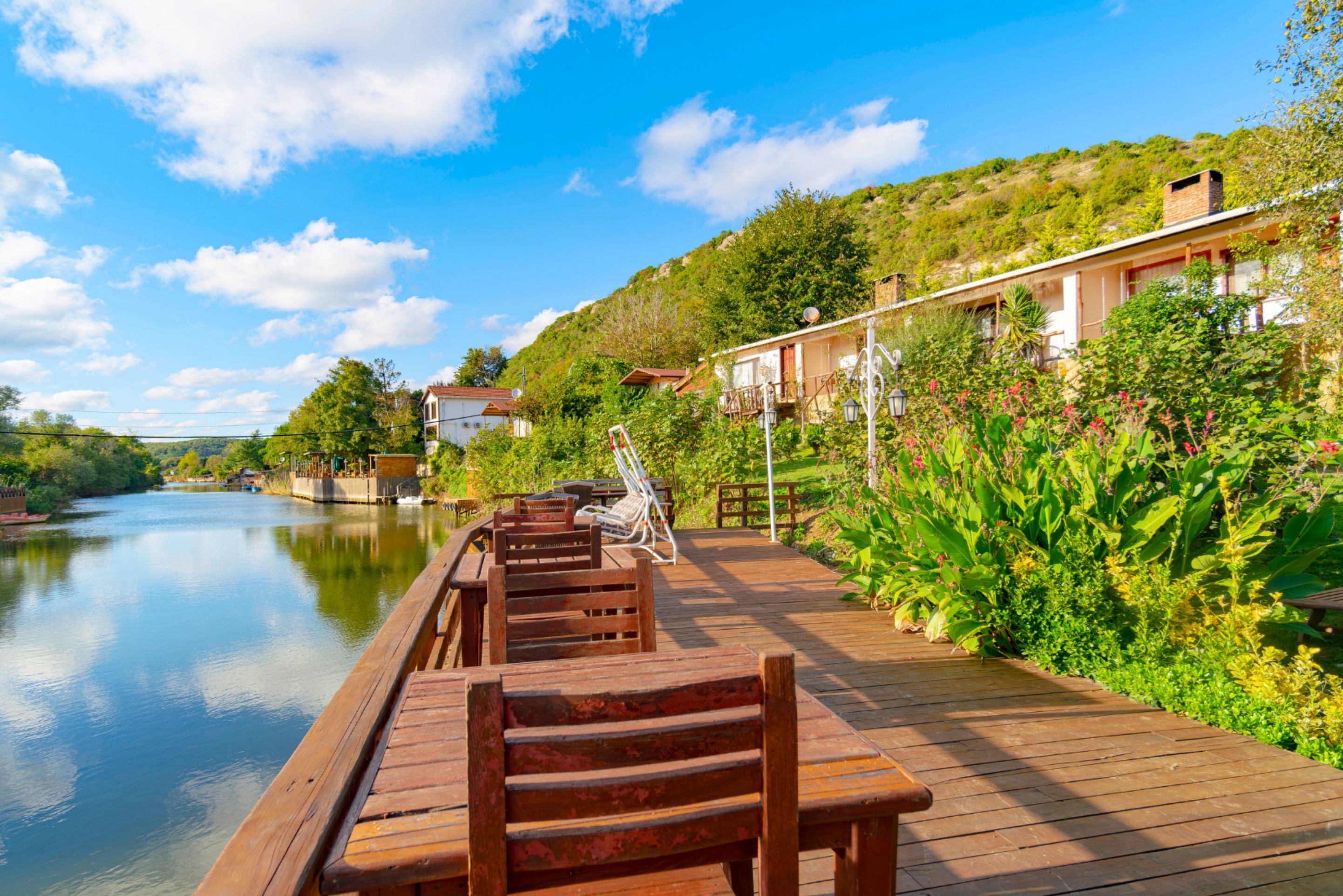 A wooden deck with benches overlooking a calm river, surrounded by lush greenery and a hillside with buildings.