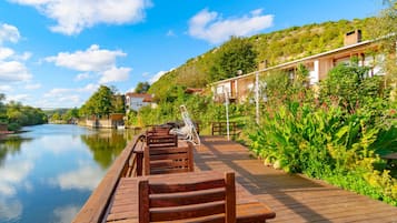 A wooden deck with benches overlooking a calm river, surrounded by lush greenery and a hillside with buildings.