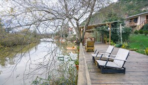 Una terraza de madera con mobiliario exterior, un estanque y un edificio con balcón al fondo.